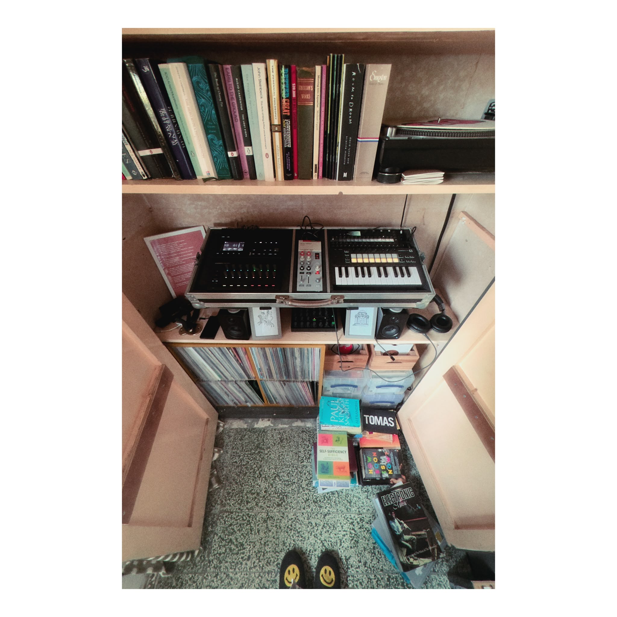A shelf of books above a flight case of synthesisers and a keyboard, studio
monitors and headphones below, a crate of records, paperbacks stacked on the
floor, and a pair of smiley-face slippers.
